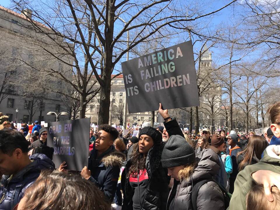 picture from the March for Our Lives. A woman holds a sign that reads "America is Failing its Children"